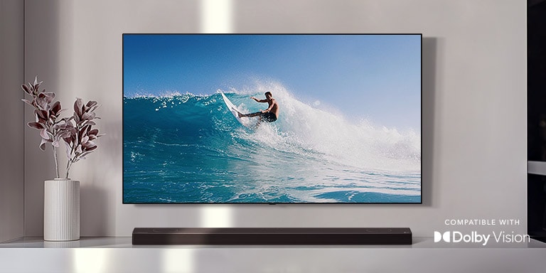 TV is on the wall. TV shows a man surfing on big wave. LG Soundbar is right below TV on a white shelf. There is a vase with a flower right next to the soundbar. (play the video) 