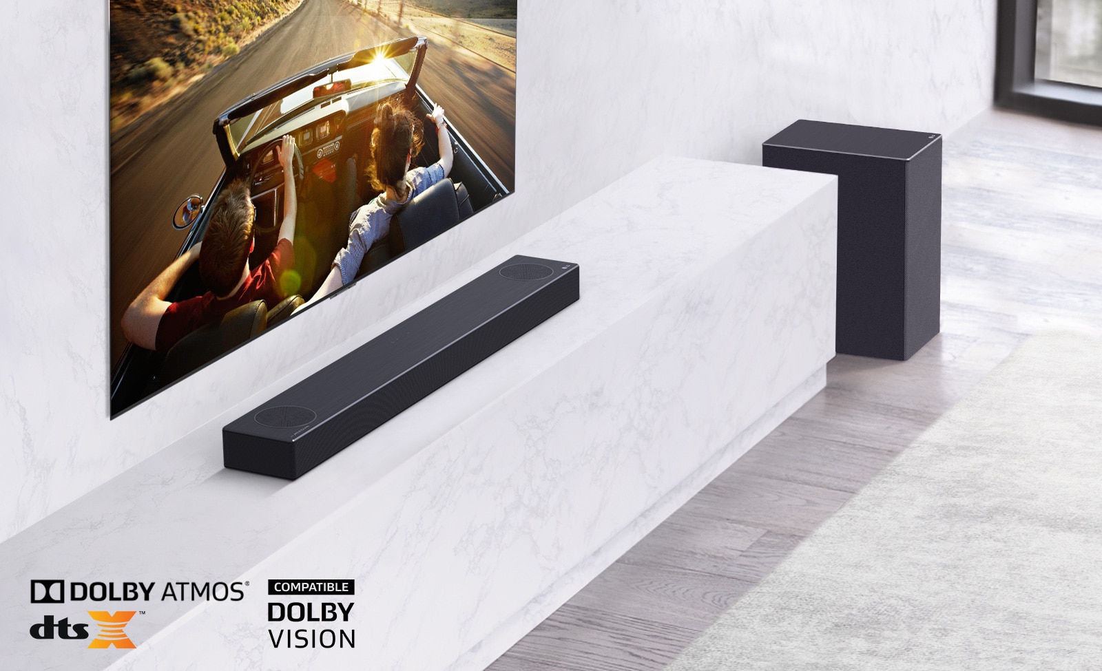 TV is on the wall, LG Soundbar is below on a white marble shelf with a sub-woofer to the right. TV shows a couple in a car. 