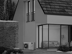 An outdoor unit located between a snow-covered brick wall and a two-storied house with wide transparent windows showing through a living room.