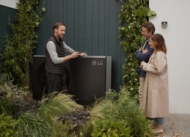 An installer in front of the LG Therma V heat pump placed on the exterior of a house is facing the couple and explaining the heating solution.