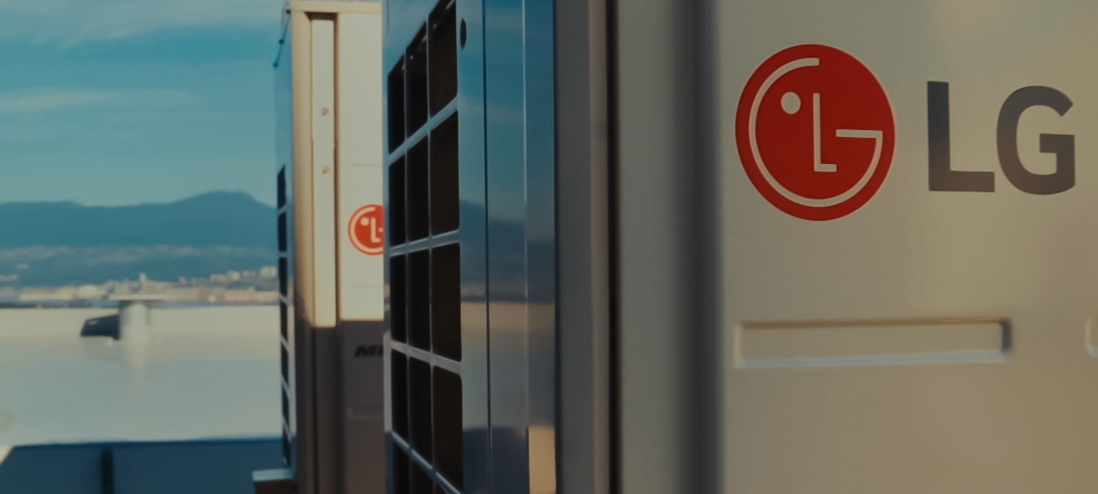 There are several LG HVAC products on the rooftop of a high-rise building. There is a mountain behind the products.