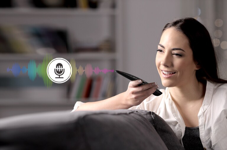 A woman is controlling TV by speaking to a remote control which has voice recognition.