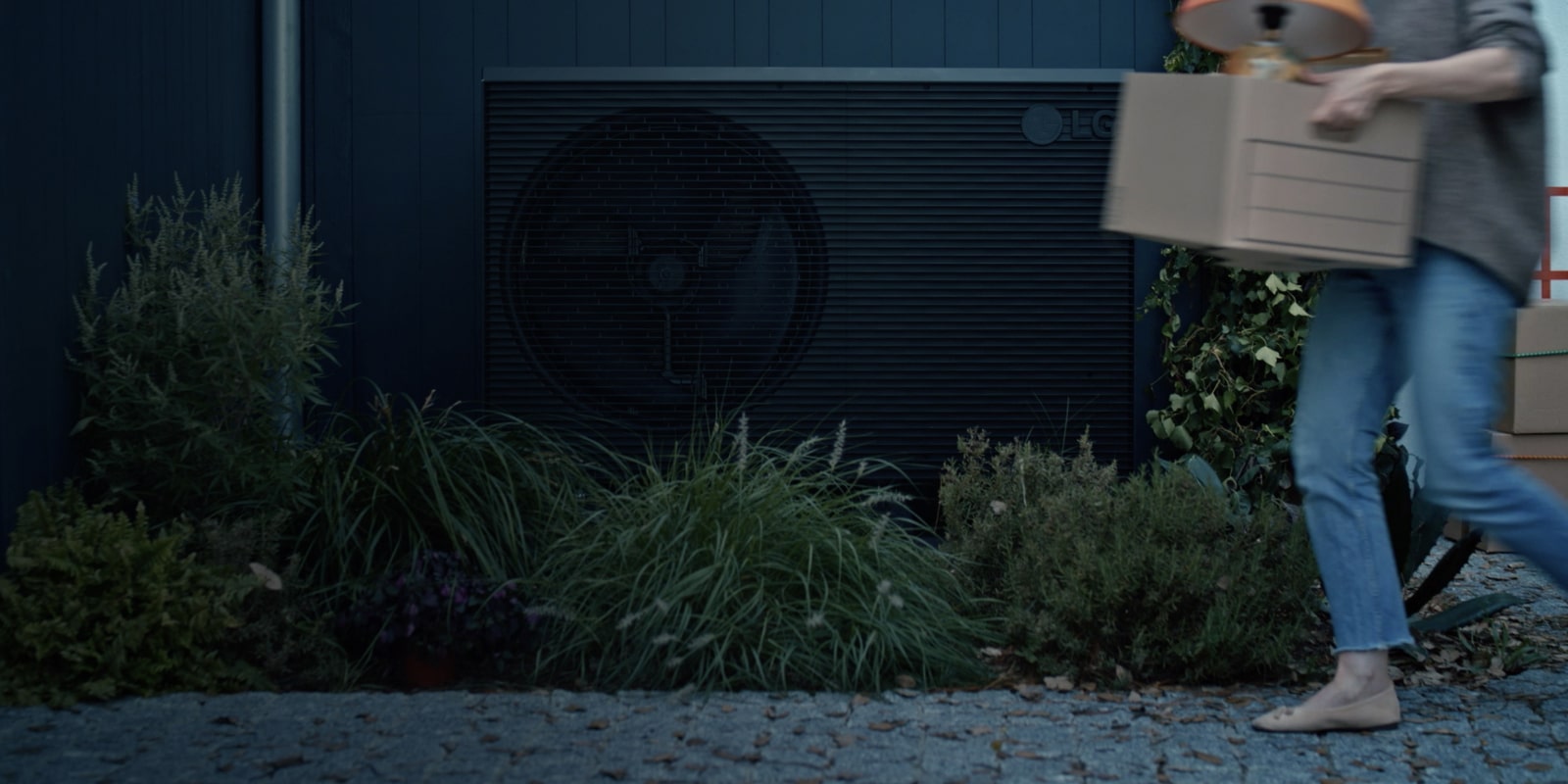A black LG THERMA V product is placed in the garden of the house and a woman with a box is passing by in front of it.