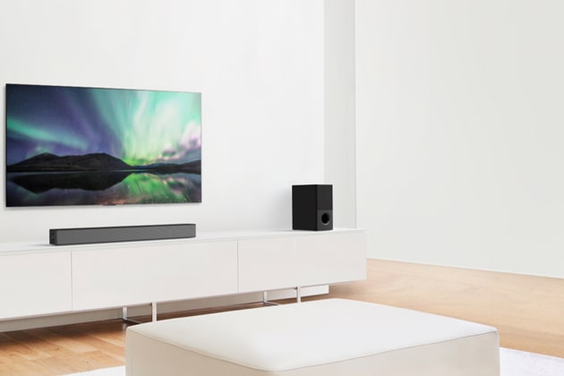 TV and sound bar in white living room with white sofa in the center. Speakers sit on both ends of the sofa.