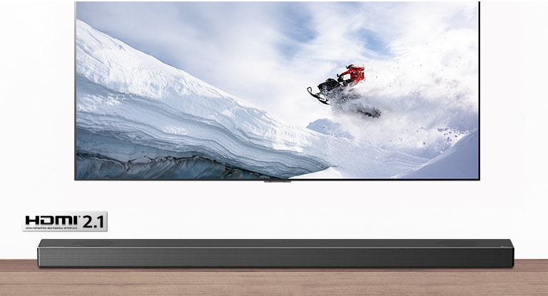 TV and Soundbar are seen from the front. TV shows man riding snowmobile in the snowy mountains. HDMI 2.1 logo is below TV. 