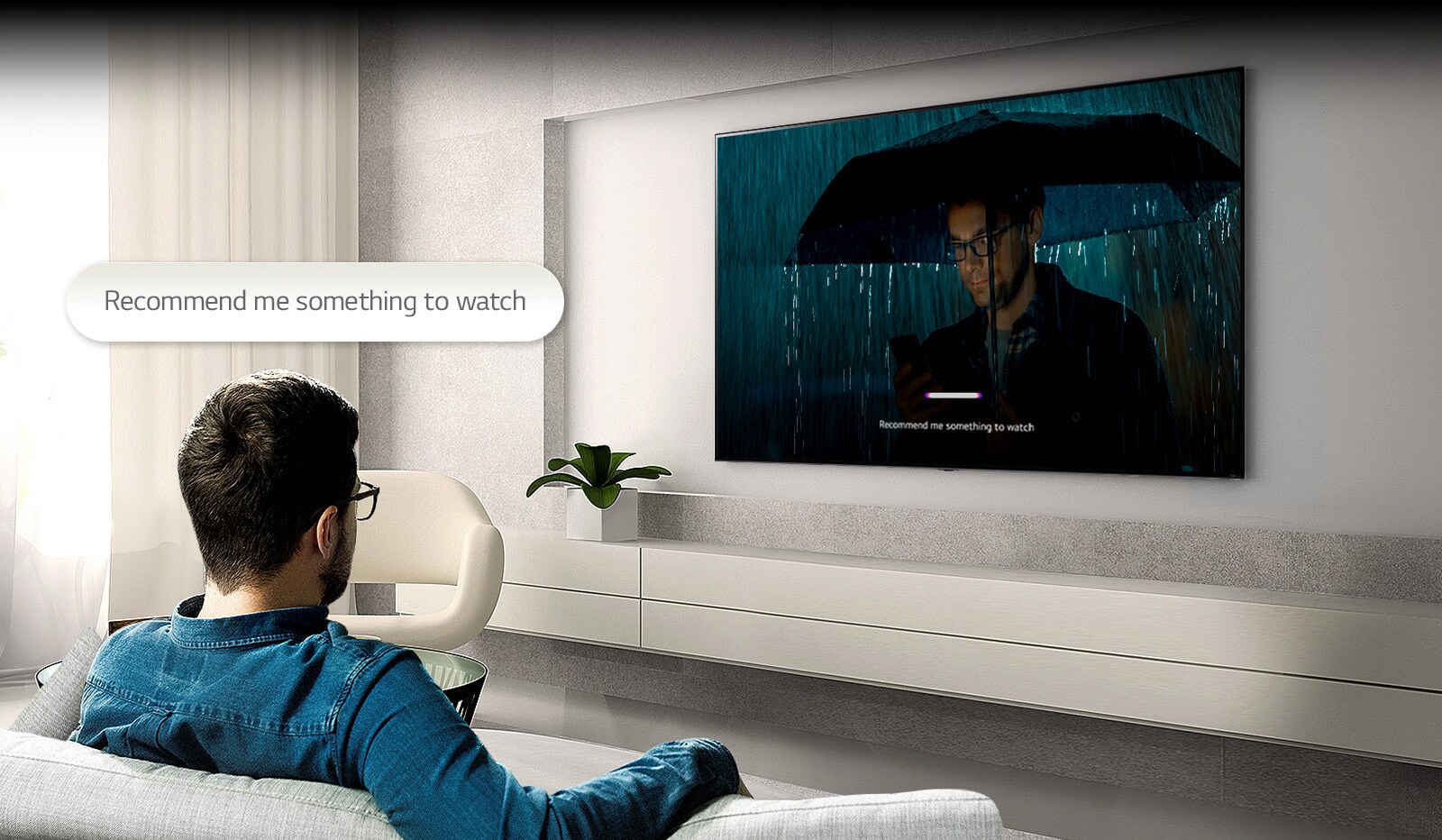 A man is sitting on a sofa facing a TV on the opposite side. A speech bubble that reads 'Recommend me something to watch' is floating above his head. Inside the screen, a video of a man wearing an umbrella is playing with a UI that is voice-recognized.