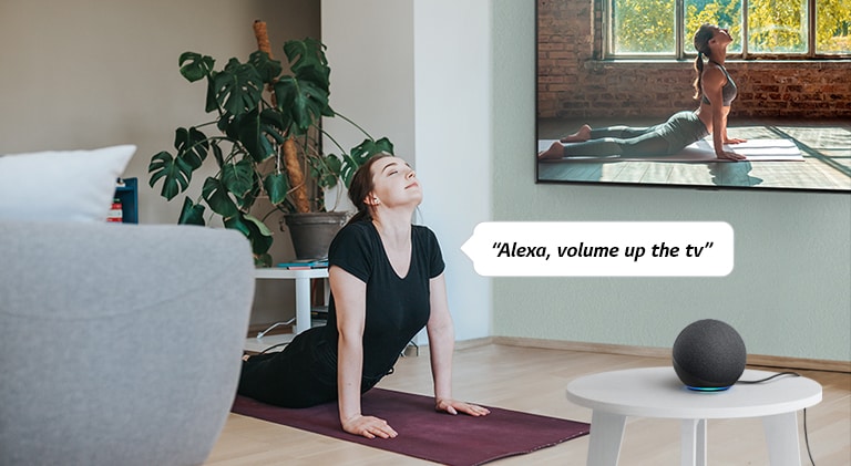 A woman following the yoga video in the living room commands an AI speaker to volume up the TV.