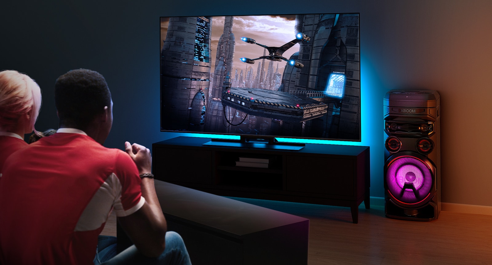 Des gens regardent la télévision sur un mur, avec une enceinte LG XBOOM RNC7 sur la droite. La télévision diffuse un film. 