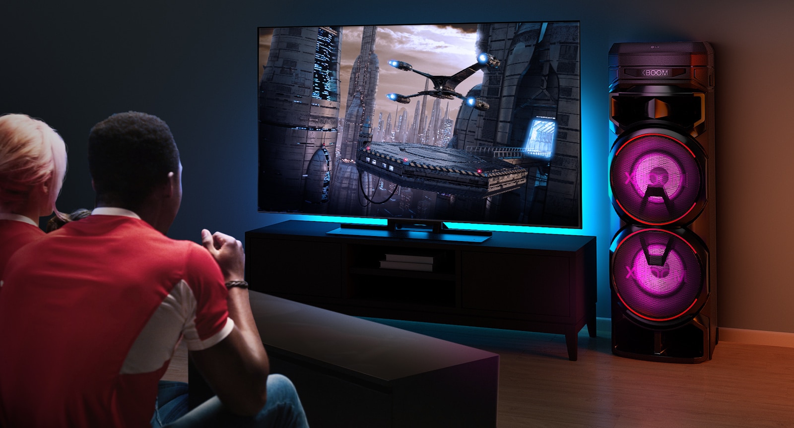 Des gens regardent la télévision sur un mur, avec une enceinte LG XBOOM RNC9 sur la droite. La télévision diffuse un film. 