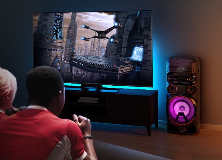 Des gens regardent la télévision sur un mur, avec une enceinte LG XBOOM RNC7 sur la droite. La télévision diffuse un film. 
