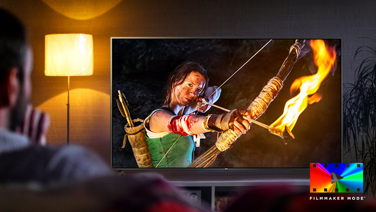 Un homme assis sur un canapé, regardant un film d’action. La fille à la télévision tient un arc complètement tendu et des flèches.