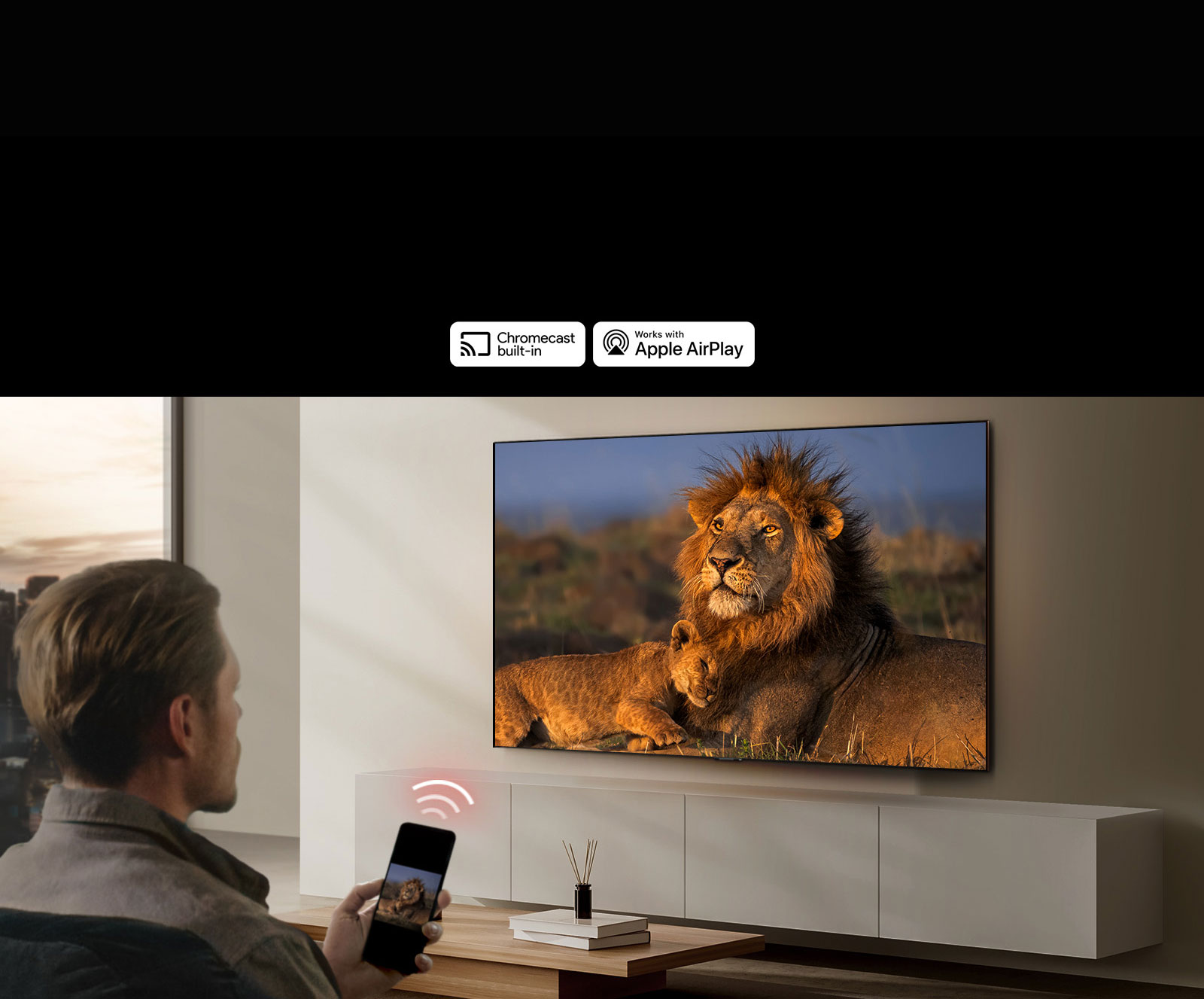  Un televisor LG montado en la pared de una sala de estar, que muestra un león y un cachorro de león. Un hombre se sienta en primer plano con un teléfono inteligente en la mano que muestra la misma imagen de leones. Un gráfico de tres barras curvas de color rojo neón se muestra justo encima del teléfono inteligente que apunta hacia el televisor.