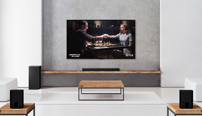 Un ensemble de 2 haut-parleurs arrière, un caisson de basses, une barre de son et un téléviseur se trouvent dans un salon blanc. Une femme et un homme jouent aux échecs sur un écran de télévision. 