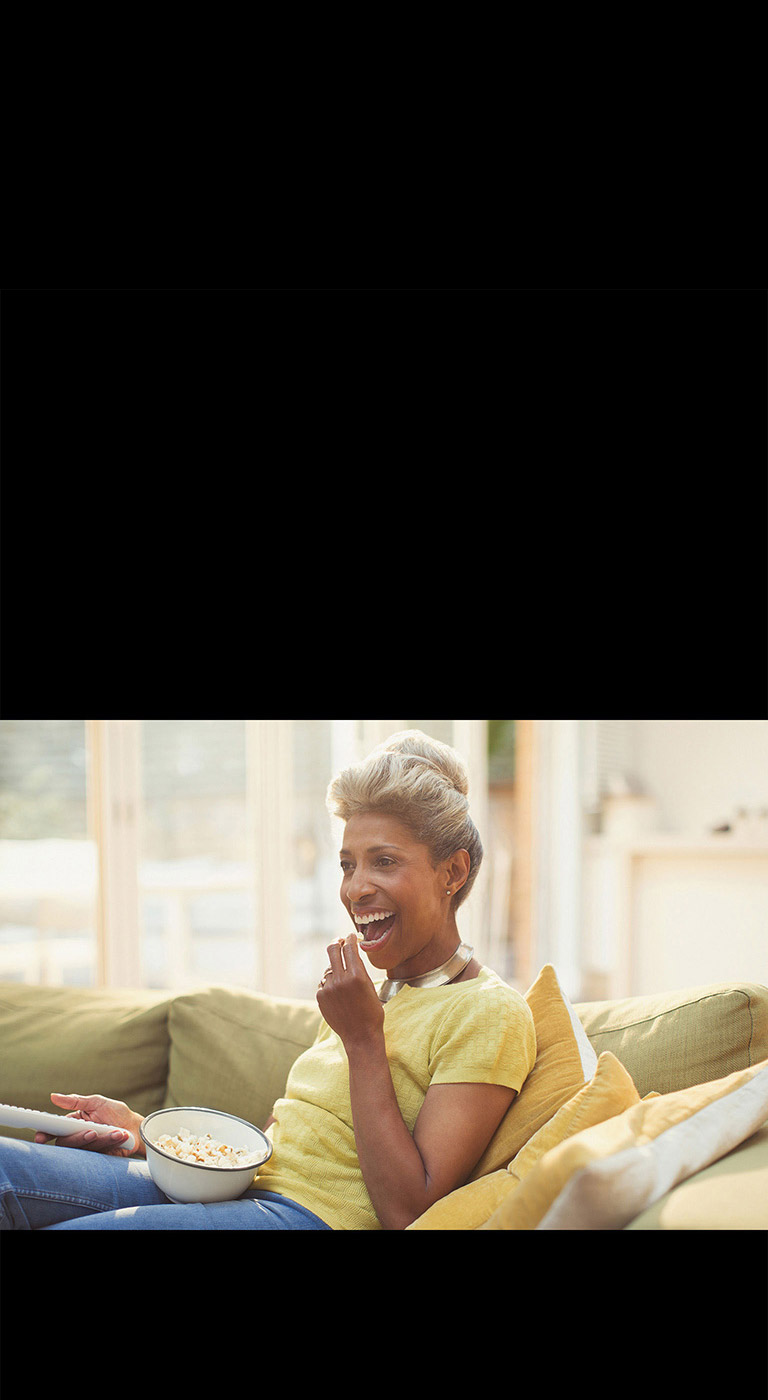 Une femme regarde le téléviseur – tenant une télécommande. Elle mange également du pop-corn.