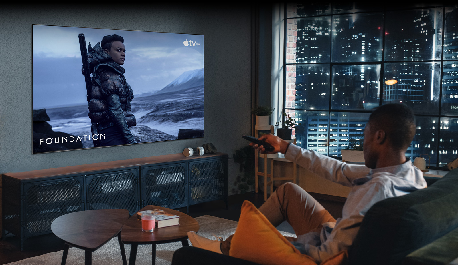 Un homme est assis sur un canapé et regarde la TV. L'homme tient une télécommande à la main et le téléviseur affiche une scène de la série Apple TV+ «&nbsp;Foundation&nbsp;».
