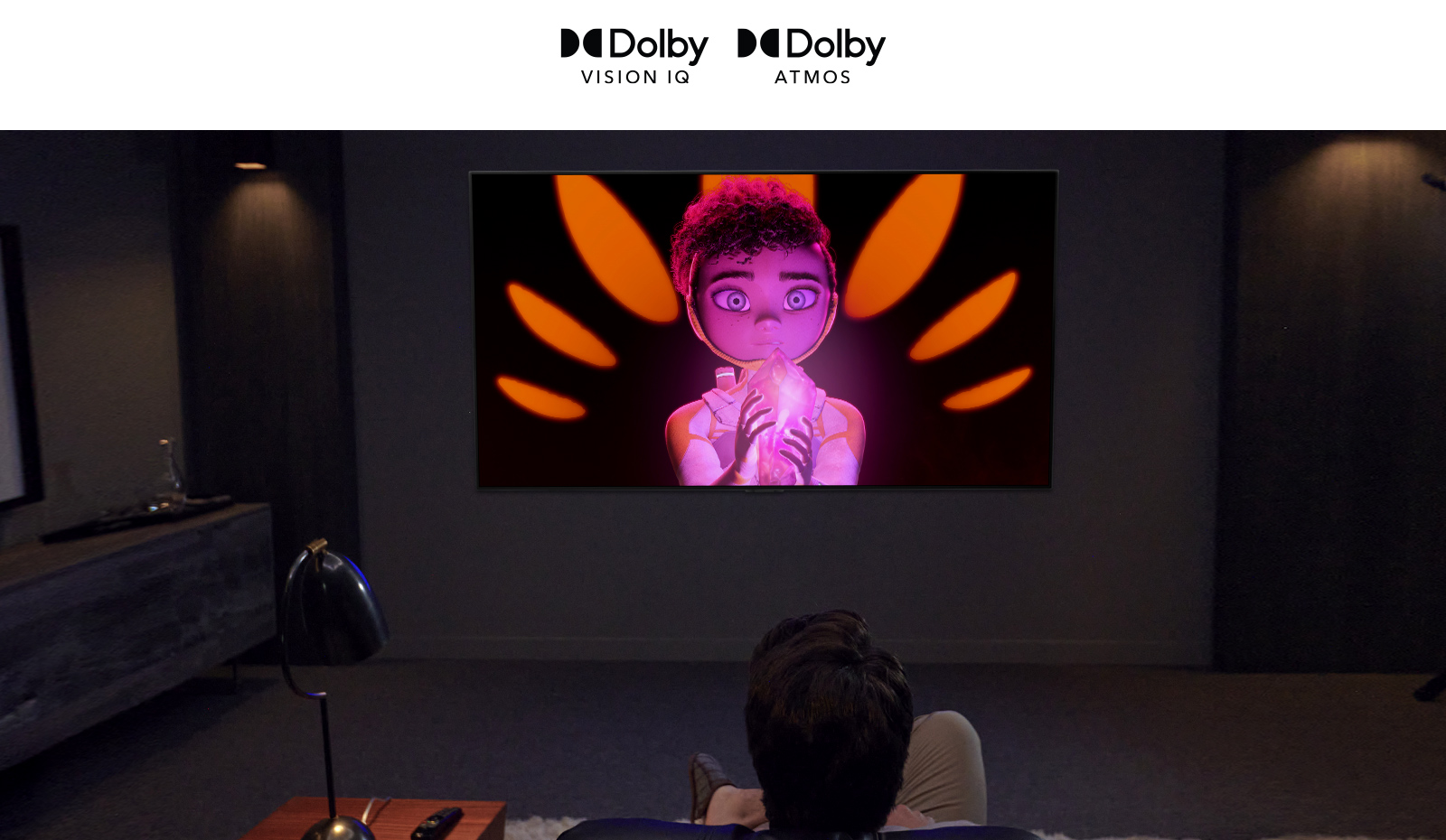 Le logo de Dolby Vision IQ et d’Atmos sont alignés horizontalement. Sous les logos, un père et son fils sont assis sur un canapé et regarde un téléviseur affichant une fille tenant un minérai au milieu d’un arrière-plan noir et orange.