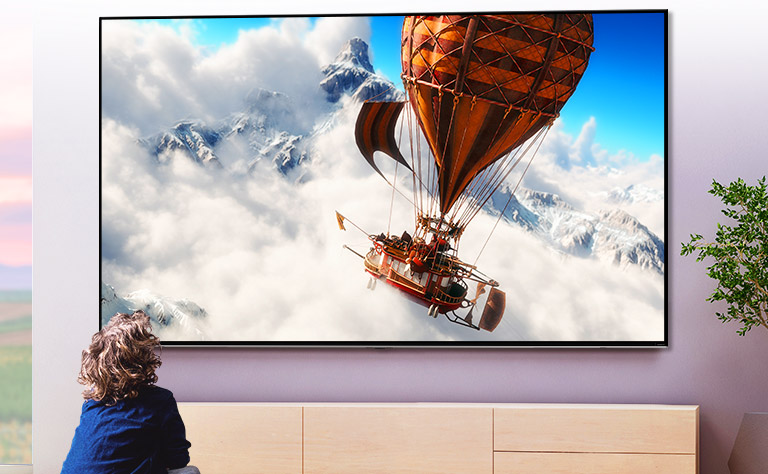 Un enfant assis devant un grand téléviseur fixé au mur dans une pièce lumineuse. L’écran affiche une montgolfière volant au milieu des nuages devant une montagne.