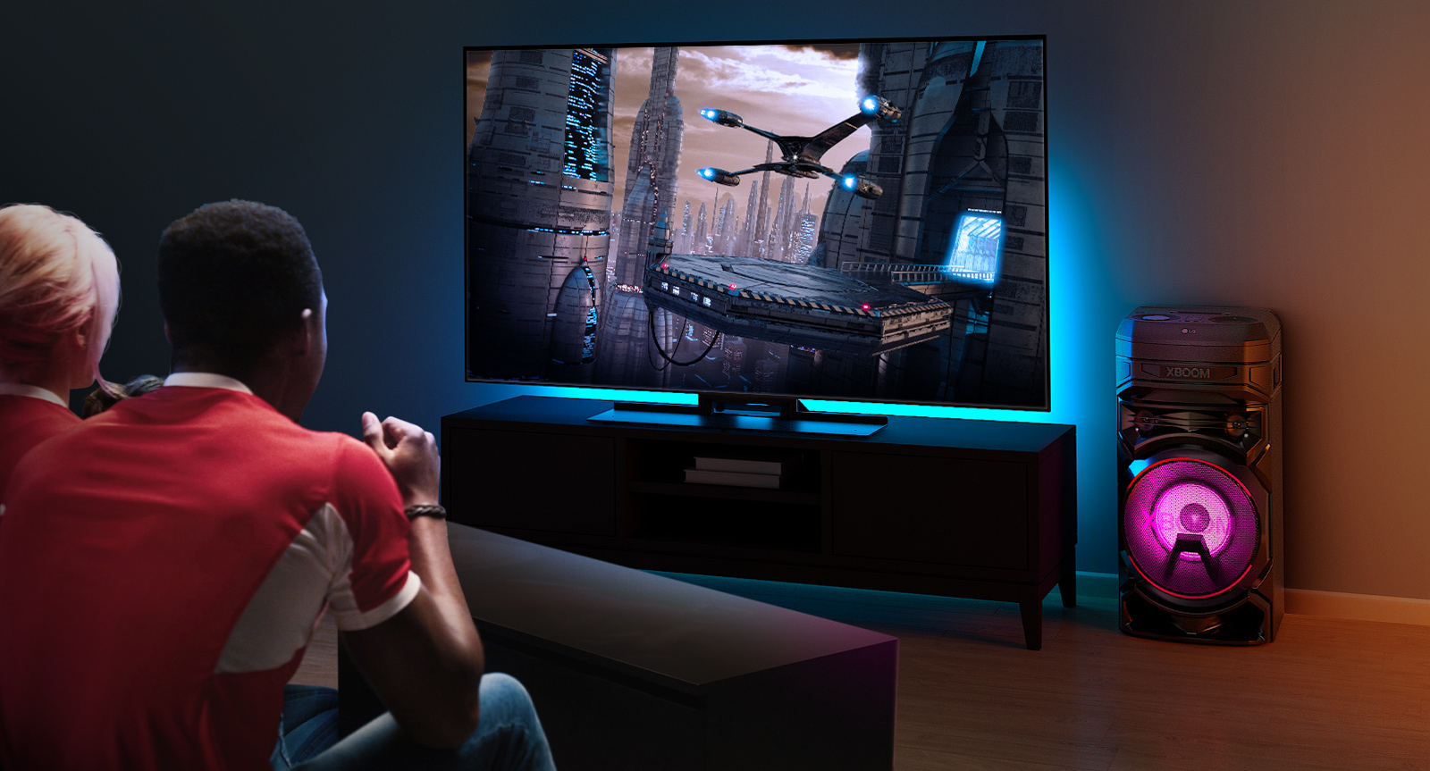 Des gens regardent la télévision sur un mur, avec une enceinte LG XBOOM RNC5 sur la droite. La télévision diffuse un film. 