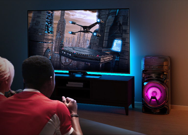 Des gens regardent la télévision sur un mur, avec une enceinte LG XBOOM RNC5 sur la droite. La télévision diffuse un film. 