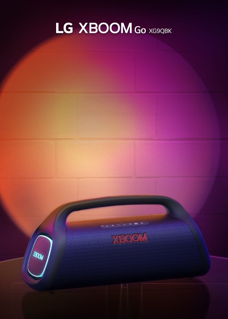LG XBOOM Go XG9 posée sur une table métallique, émet une lumière orange. Derrière la table, l’éclairage scénique illumine le mur