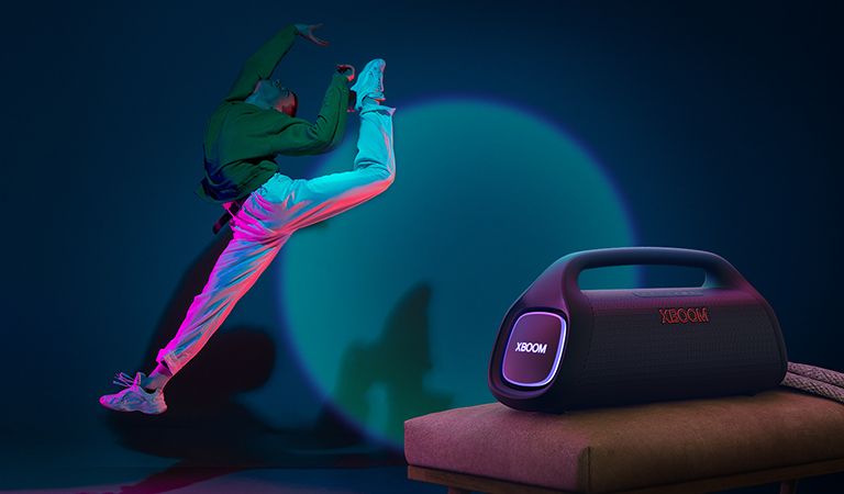 La LG XBOOM Go XG9 est posée sur la chaise. Elle émet une lumière scénique sur le mur et une personne danse devant.