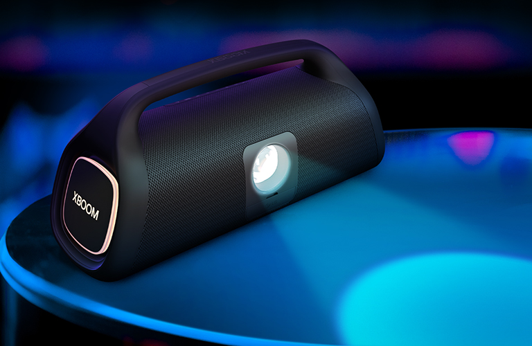 La LG XBOOM Go XG9 est posée sur la surface, montrant son ampoule d’éclairage scénique. L’ampoule projette une lumière bleue, et sur le côté, un éclairage orange est visible.