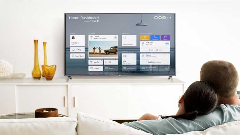 Un homme et une femme assis sur un canapé dans le salon et regardant le tableau de bord Accueil affiché à l’écran du téléviseur.