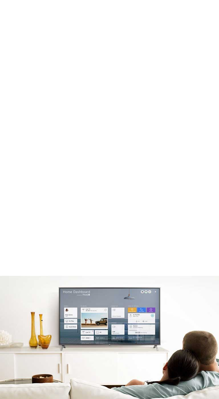 Un homme et une femme assis sur un canapé dans le salon et regardant le tableau de bord Accueil affiché à l’écran du téléviseur.