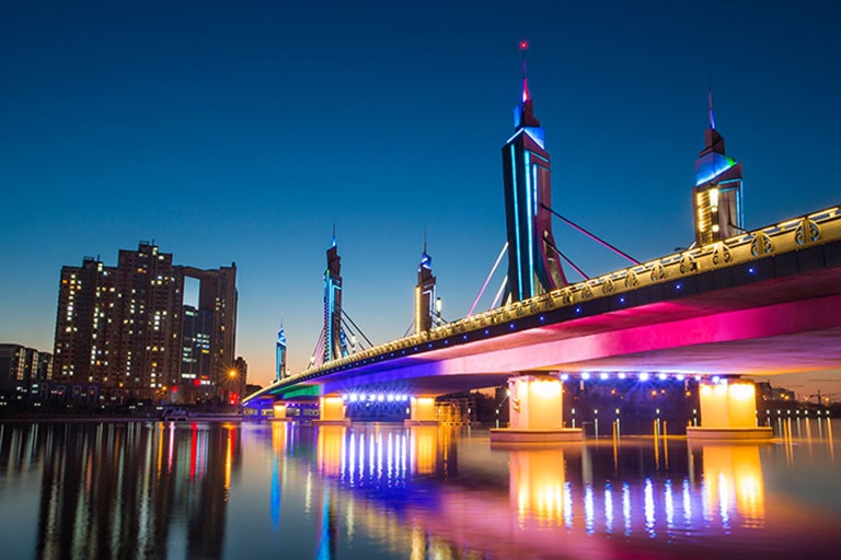 Vista nocturna de un puente iluminado con rascacielos de fondo y luces vívidas reflejadas en el agua, para demostrar el alto contraste y el detalle del color.