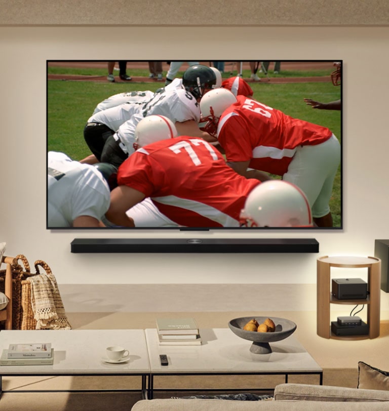 Una sala de estar con una LG True Wireless TV en la pared. En la pantalla se ve un partido de fútbol americano. La instalación de la TV también incluye una LG Soundbar montada en la pared. Y, casi imperceptibles, el resto de los dispositivos periféricos están conectados a una Caja de conexión cero que se encuentra sobre una mesa de apoyo. La sala de estar está prolija, ordenada y no hay desorden de cables.