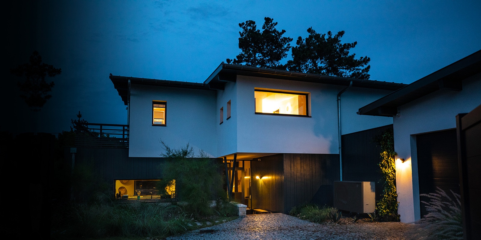 En una noche fría de invierno, plano general del exterior de la casa suavemente iluminada en el interior. Colocación de la nueva bomba de aire-agua negra THERMA V de LG en frente de la casa. 