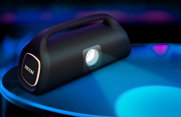 LG XBOOM Go XG9 se coloca en la superficie, mostrando su bombilla de iluminación de escenario. La bombilla proyecta un foco azul y, en el lateral, hay una iluminación orgánica encendida.