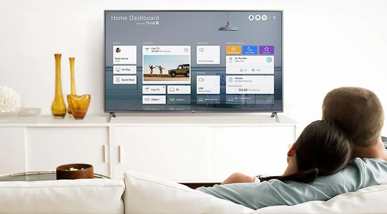 Un hombre y una mujer sentados en un sofá en la sala de estar con el Panel para el hogar en la pantalla del televisor