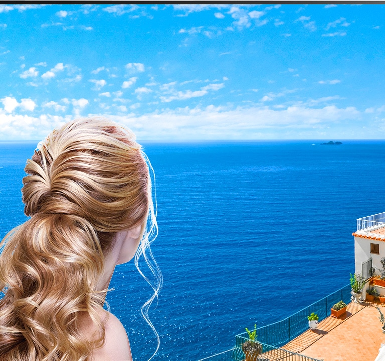 Vista trasera de una mujer mirando por la ventana un mar azul y edificaciones de color blanco. Al deslizarse en la página hacia abajo, la imagen se aleja para mostrar más de la habitación y revelar que la ventana es un televisor LG QNED Mini LED.