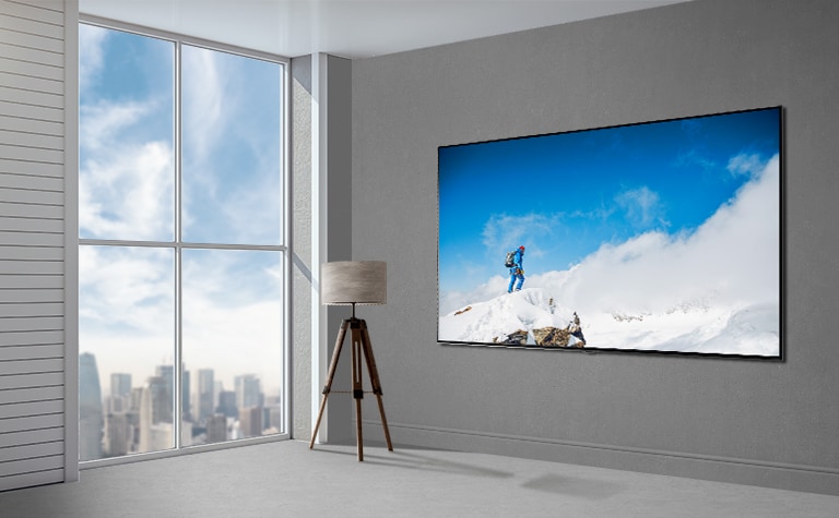 La imagen de un televisor de pantalla plana montado en una pared gris junto a una ventana que va del suelo al techo muestra un paisaje urbano. Hay una lámpara a la izquierda del televisor y la pantalla muestra a un excursionista en una roca cubierta de nieve con un cielo azul y nubes de fondo.