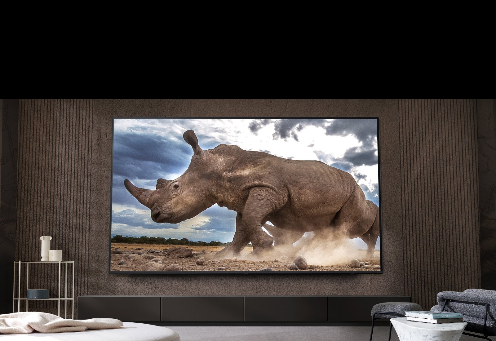 Auf einem Ultra Big LG Fernseher, der an der braunen Wand eines Wohnzimmers montiert ist, umgeben von cremefarbenen modularen Möbeln, wird ein Nashorn in einer Safari-Umgebung gezeigt.