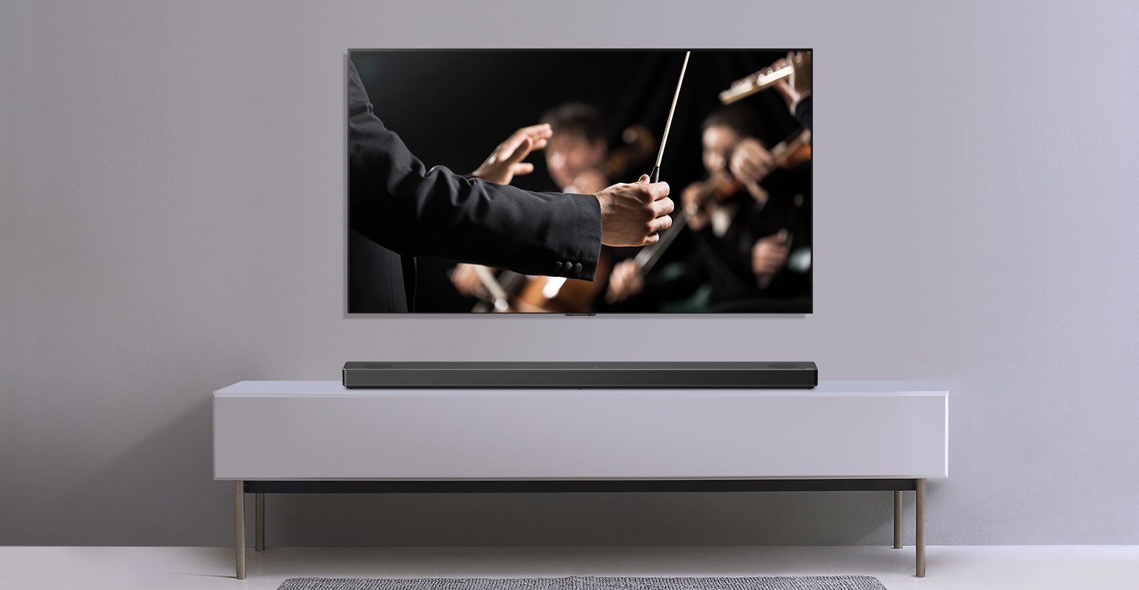 Un televisore è mostrato davanti a un muro grigio e una LG Sound Bar sotto su uno scaffale grigio. La televisione mostra un direttore che dirige un'orchestra.