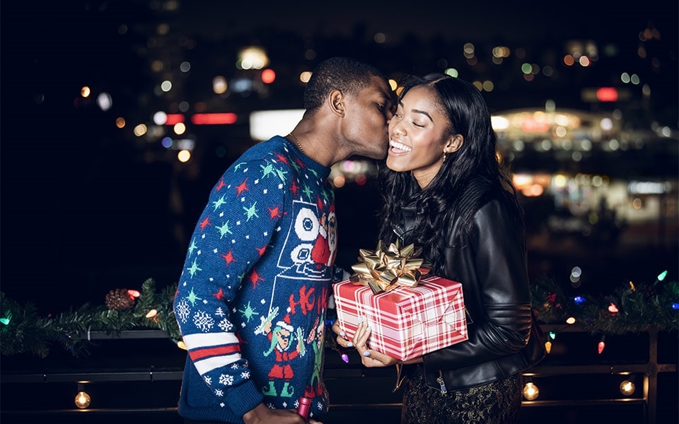 A smiling woman holds a Christmas present while her partner kisses her on the cheek
