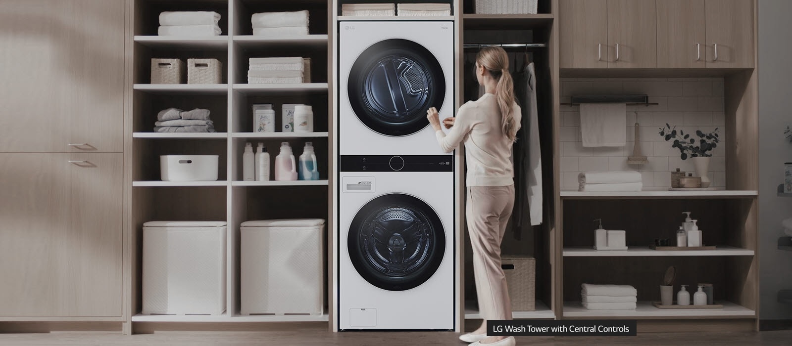 A white washer and dryer are stacked on top of each other and installed in a wall unit with shelves and a closet and a woman has her back facing the front as she reaches up to press a button on the center top control panel. She stands uncomfortably.