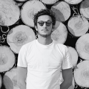 A black and white head-on shot of Delfino Sisto Legnani stood in front of stacks of timber.