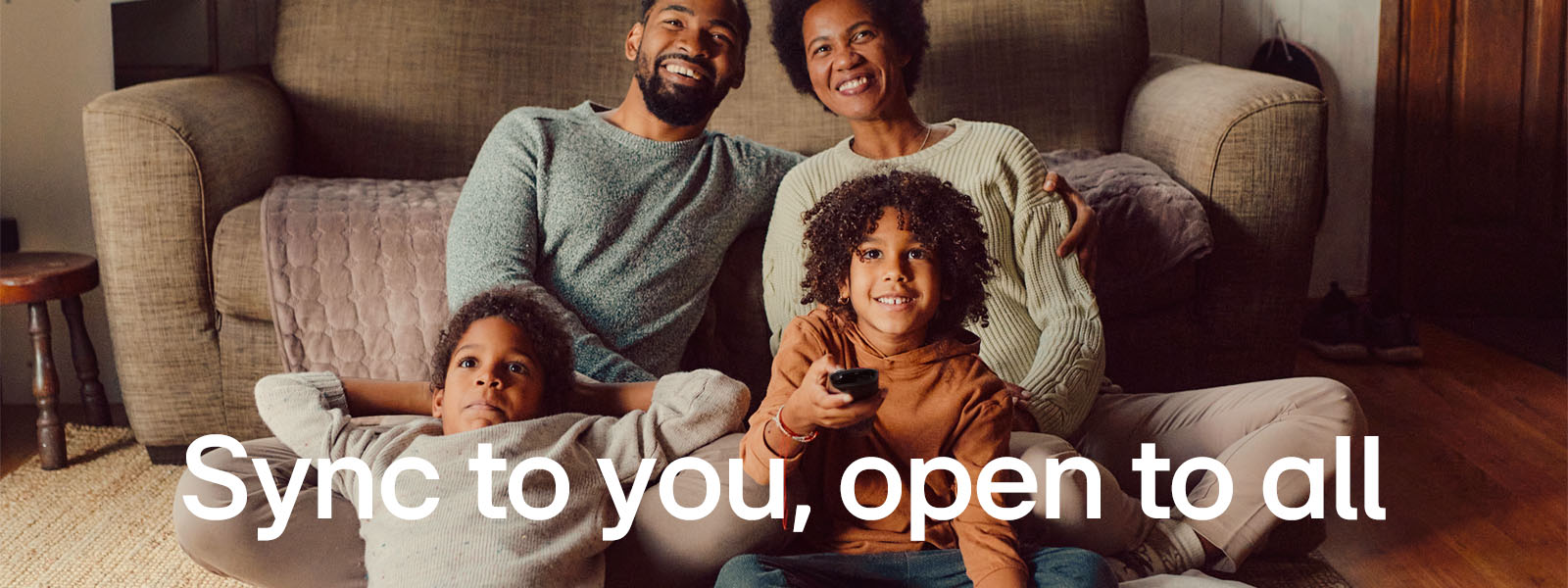 An image of a family of four sitting on the floor in front of cozy brown sofa looking straight ahead and laughing while watching TV. The words "Syncs to you, open to all" overlay the image in a white font. 