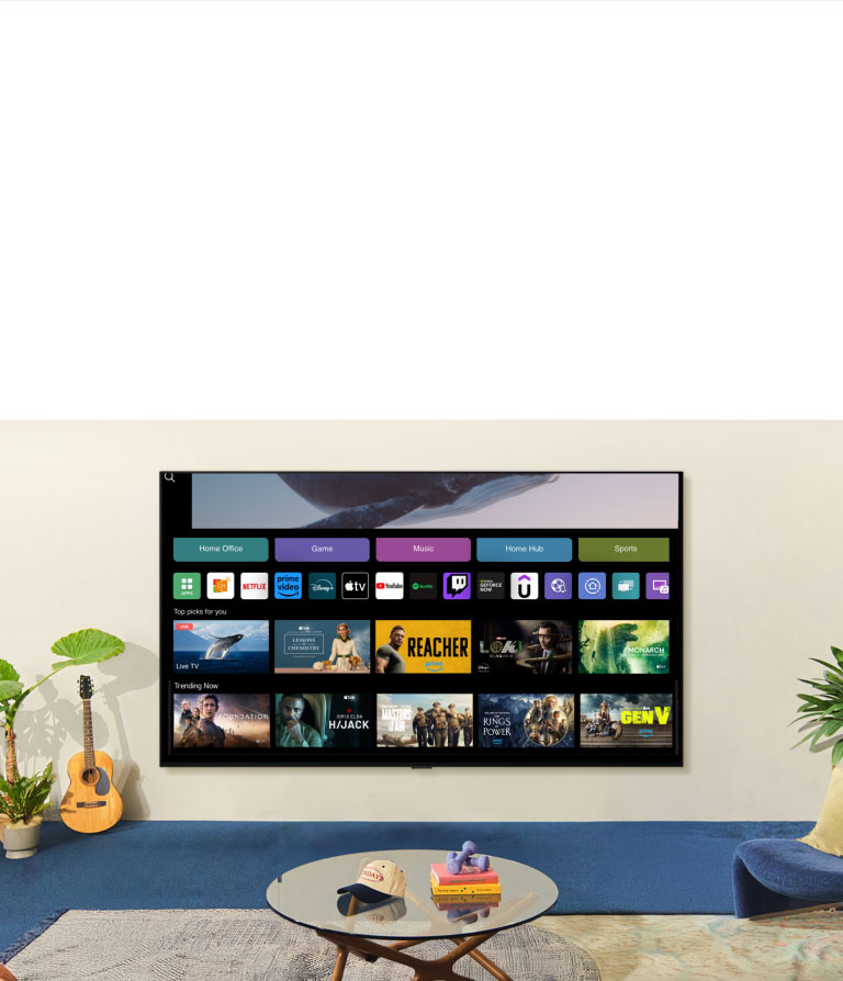 A whale floating above the ocean with a woman in the ocean are shown. A home screen appears from below. As the scene transitions, it shows the image in a large LG TV on the wall. Two women sit in a cozy and neutral living room filled with plants, and a guitar. One woman points the remote at the TV, which shows a range of apps and recommended TV shows on the home screen.