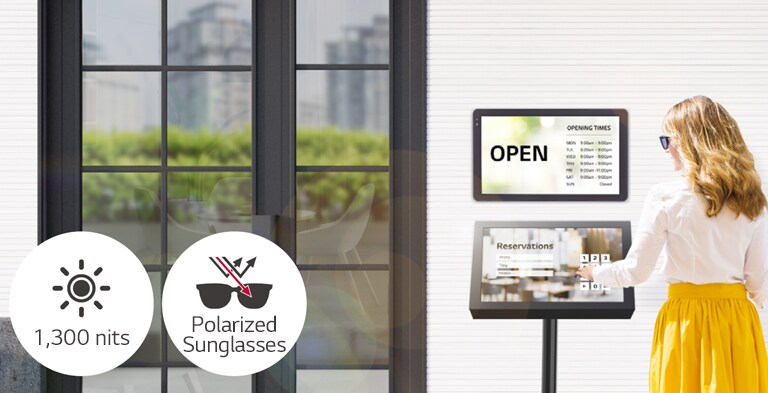 A 22XF1TJ is installed on the wall at the entrance of the building with another 22XF1TJ stand below for reservation purposes. The lady with sunglasses is using the touchscreen to make a reservation. The screen remains visible even when under direct sunlight.