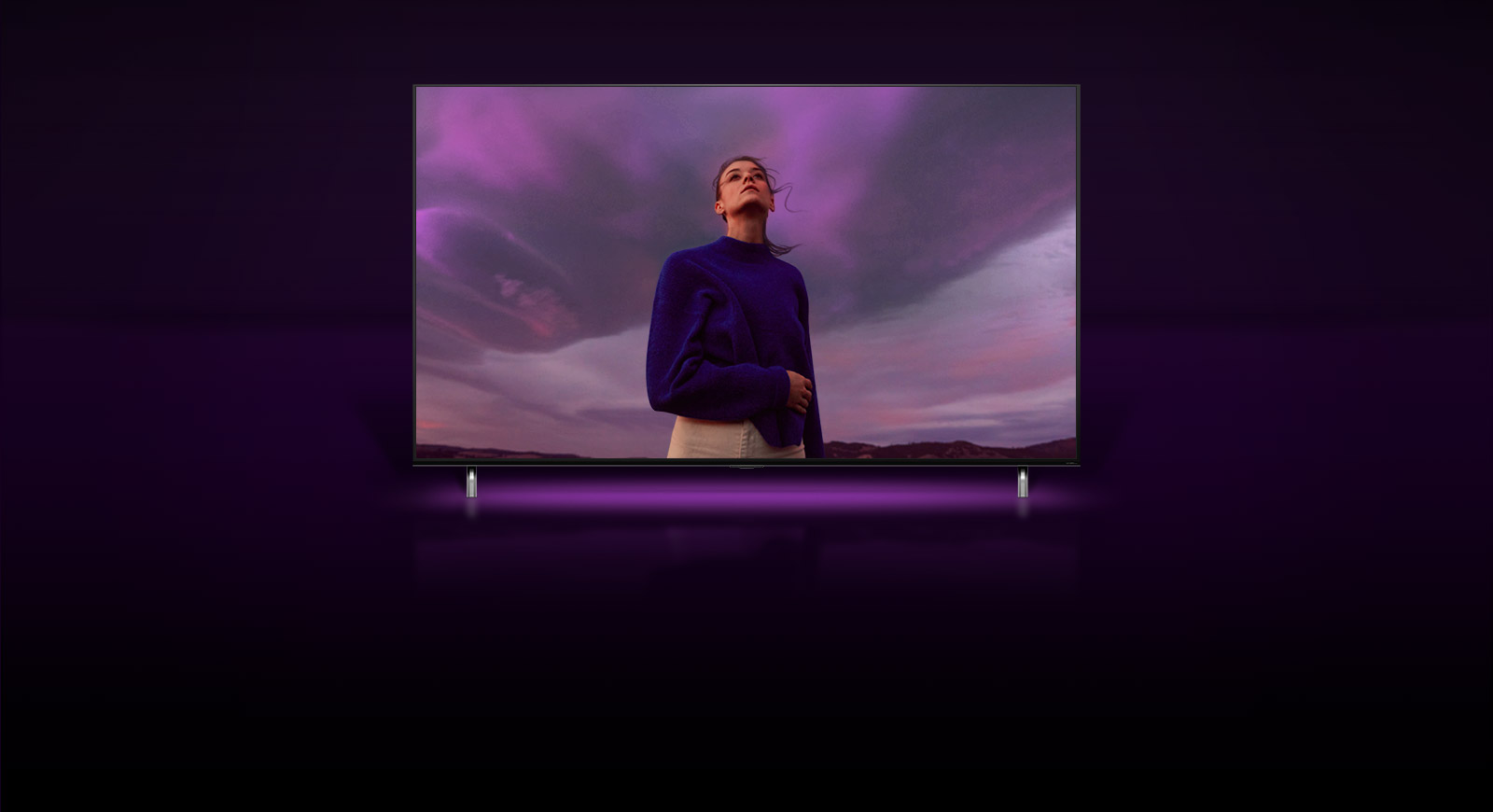 A woman is standing looking at a purple sky and the image zooms out to show the same woman in QNED TV screen.