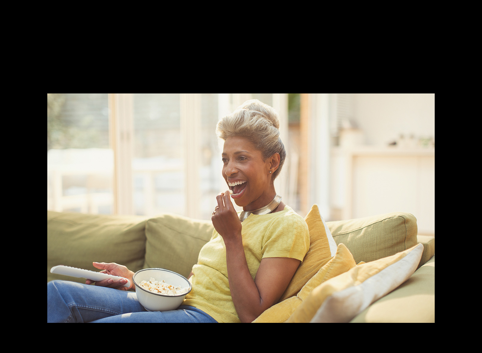 A woman is watching TV – holding a remote controller. She is also eating popcorn.