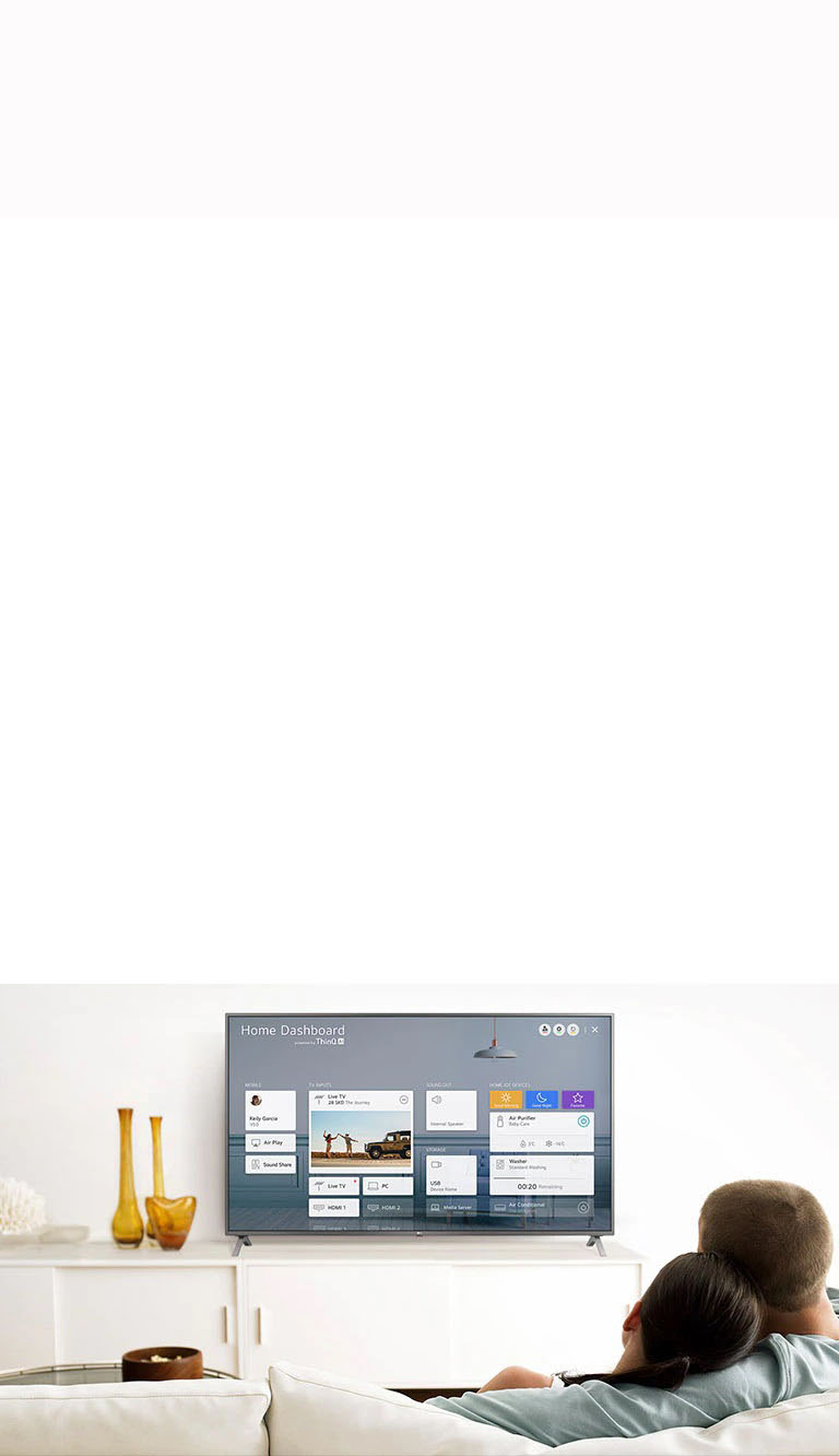 A men and women sitting on a sofa in the living room with the Home Dashboard on the TV screen.