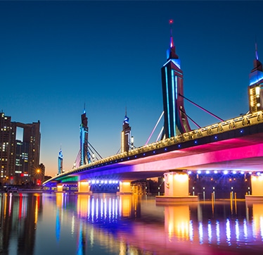 Vue nocturne d’un pont moderne et de gratte-ciels illuminés avec un éclairage vif qui se reflète dans l’eau, démontrant un contraste élevé et les détails des couleurs.
