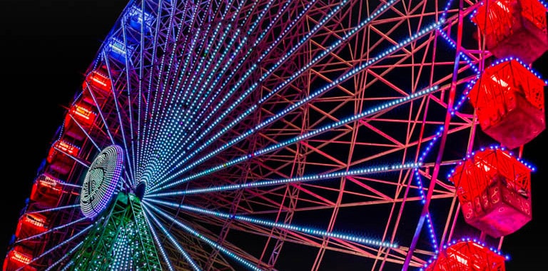 Une grande roue légèrement décolorée s’illumine la nuit, avec des éclats colorés partant du côté gauche de l’écran et se déplaçant sur celui-ci. Au fur et à mesure que les étincelles colorées défilent, l’écran passe d’un voile nuageux à des couleurs éclatantes et lumineuses.
