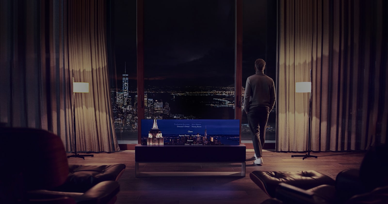 Un homme tourné vers l'arrière observe la vue nocturne de la ville à travers sa grande fenêtre avec le téléviseur OLED enroulable LG SIGNATURE en ligne derrière lui.