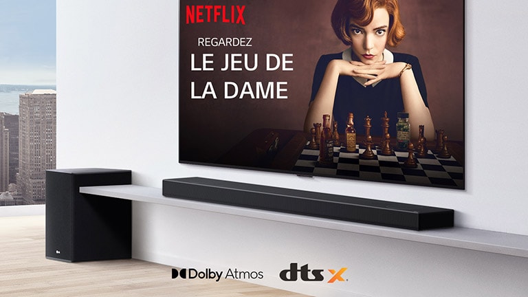 Le téléviseur est fixé au mur. Une affiche d’une émission de télévision est diffusée sur l’écran du téléviseur. La barre de son LG se trouve juste en dessous du téléviseur sur une étagère blanche avec un caisson de basses juste à côté. Logos Dolby Atmos et DTS: X affichés au milieu en bas de l’image.